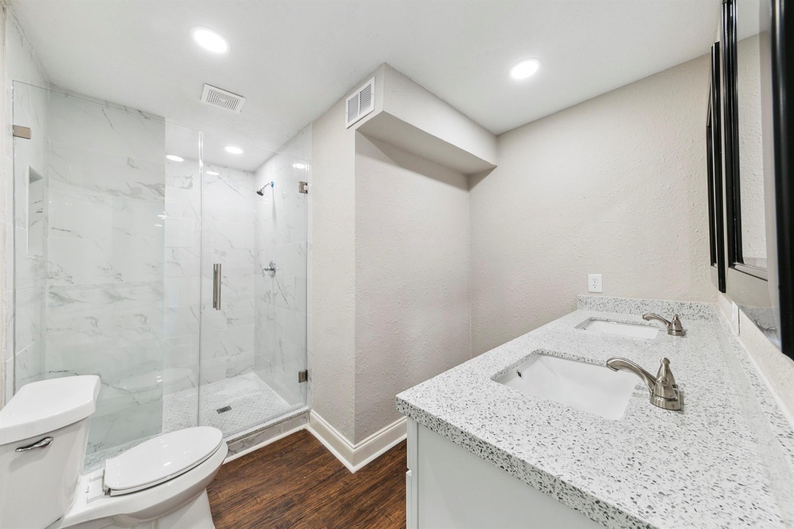 Completed bathroom with glass shower, dual vanity, stone-look counters, and wood-look flooring